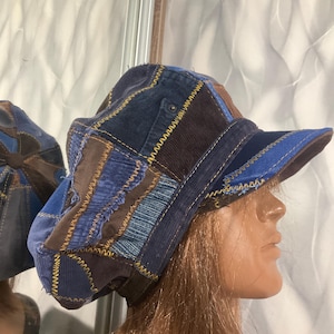Puede incluir: Una gorra de patchwork construida con varios paneles de tela azul y marrón, incluyendo texturas de pana y mezclilla. Las piezas están unidas con costuras en zigzag amarillas visibles, con una visera estructurada y forma redondeada.