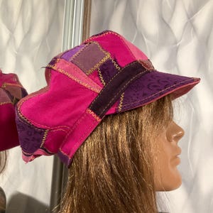 Puede incluir: Una gorra tipo boina de patchwork con paneles de tela en varios tonos de rosa brillante y morado oscuro. El gorro presenta costuras en zigzag amarillas visibles a lo largo de las uniones y una visera estructurada de tela morada con estampado.