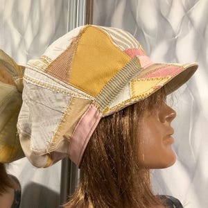 May include: A patchwork cap constructed from various fabric panels in shades of yellow, pink, beige, and white. The hat features visible frayed edges, decorative zigzag stitching, and a structured brim, showcasing a multi-textured design.