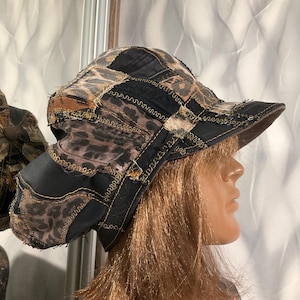 Puede incluir: Un sombrero de pescador de estilo patchwork que combina paneles de tela negra sólida con paneles de tela con estampado de leopardo en tonos marrones y negros. El sombrero presenta bordes deshilachados y costuras decorativas en zigzag.