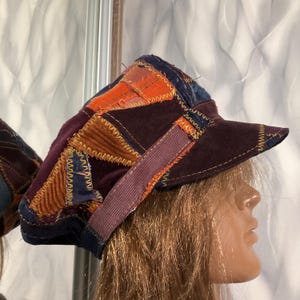 Puede incluir: Una gorra de estilo newsboy hecha de parches con diversas texturas y colores de tela, incluyendo morado oscuro, naranja y azul. Los paneles están unidos con costuras en zigzag visibles y la gorra incluye una visera y una banda ancha.