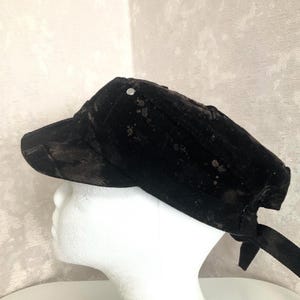 May include: A black military-style cap featuring a subtle, dark mottled or paint-splatter pattern across the fabric. The cap has a structured brim, a single silver-toned metal stud detail on the side, and fabric ties at the back for adjustment.