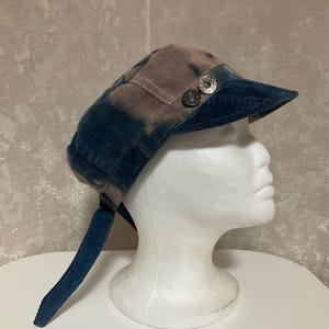 May include: A corduroy military-style cap featuring a tie-dye pattern in shades of blue and brown. The cap includes a short brim, a long fabric strap hanging from the side, and two silver-toned metal buttons attached to the side of the crown.