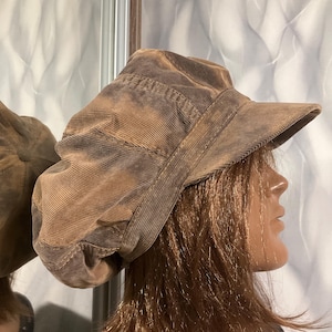 May include: A brown corduroy newsboy cap with a textured, ribbed fabric surface and a structured brim. The hat features a multi-paneled construction with visible stitching along the seams, creating a rounded, voluminous shape that sits on the head.