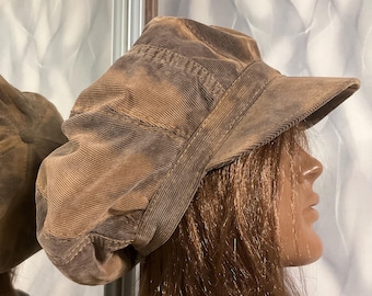 May include: A brown corduroy newsboy cap with a textured, ribbed fabric surface and a structured brim. The hat features a multi-paneled construction with visible stitching along the seams, creating a rounded, voluminous shape that sits on the head.