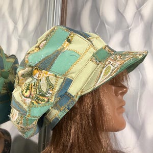 May include: A patchwork newsboy cap constructed from various fabric panels in shades of teal, light green, and yellow. The cap features a prominent paisley print on select sections, visible zigzag stitching along the seams, and a stiffened brim.