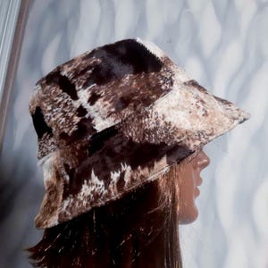 May include: A brown and white patterned bucket hat. The hat has a cowhide-like design with patches of dark brown, light brown, and white. The hat is made of a soft, textured material.