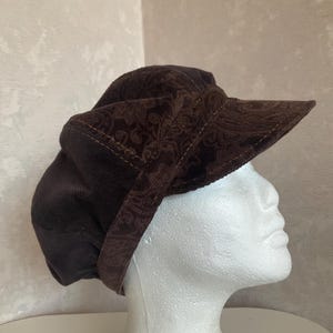 May include: A brown corduroy hat with a paisley pattern on the brim and a rounded crown. The hat has a short, stiff brim and is set against a neutral background. The hat is a dark brown color.
