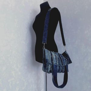 May include: A denim crossbody bag with a dark blue strap and a matching body. The bag is made from recycled denim, with visible seams and a unique texture. The bag is displayed on a black mannequin.