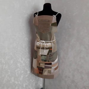 Peut inclure: Tablier patchwork sur mannequin. Il est composé de divers tissus aux tons terre, dont du denim et du kaki, avec des pièces à motifs. Il possède une poche et une attache blanche à la taille. Les bretelles sont blanches.