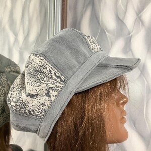 Puede incluir: Gorra de mezclilla gris con un estampado de piel de serpiente en los paneles laterales y el borde. La gorra tiene un diseño estructurado con una visera corta y un ribete cosido. Un accesorio de moda para ropa casual.
