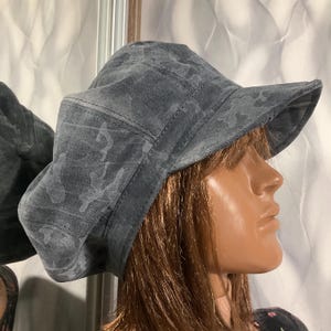 Puede incluir: Un sombrero de pescador gris con un estampado de camuflaje. El sombrero tiene un ala estructurada y una corona redondeada. La tela parece ser un material suave y duradero. El sombrero está diseñado para la protección solar y un estilo casual.