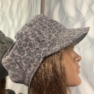 May include: A gray and black leopard print newsboy cap featuring a structured multi-panel crown and a stiff, forward-facing brim. The hat is constructed from a textured fabric with a repeating animal spot pattern across the entire surface.