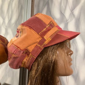 May include: A patchwork cap featuring a variety of fabric panels in shades of orange, deep red, and maroon. The cap is constructed with visible, thick, decorative stitching along the seams, connecting the different coloured fabric sections.