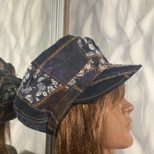 May include: A patchwork cap constructed from dark blue denim and fabric panels featuring a white floral pattern. The sections are joined with visible brown zigzag stitching, and the cap includes a stiffened brim and a rounded crown structure.