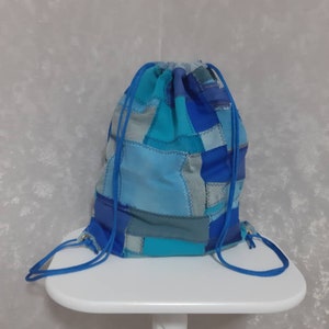 Puede incluir: Una mochila con cordón con un diseño de patchwork en tonos azules y turquesas. La bolsa presenta un cierre de cordón azul y está hecha de cuadrados de tela cosidos. La bolsa está sobre una superficie blanca.