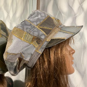 Op de afbeelding: Een patchwork hoed in tinten grijs, bruin en olijfgroen. De hoed heeft een klep en is gemaakt van verschillende stukken stof die aan elkaar zijn genaaid met goudkleurig garen. De hoed wordt getoond op een mannequinhoofd.