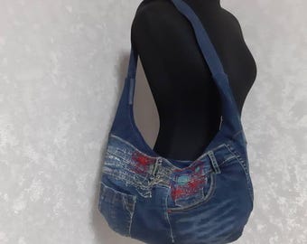 May include: A denim shoulder bag with a blue strap. The bag is made from recycled jeans, featuring pockets and red and turquoise embroidery. The bag is displayed on a black mannequin.