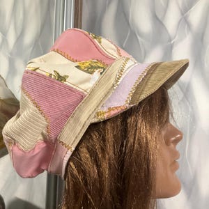 Puede incluir: Una gorra estilo boina de patchwork que combina paneles de pana en tonos rosa, crema y beige con secciones de tela con estampado floral. Las piezas están unidas mediante costuras decorativas en zigzag visibles y cuenta con una visera rígida.