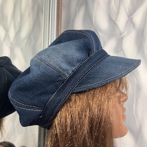 May include: A denim newsboy cap with a short brim and a band around the crown. The hat is a medium blue color with tan stitching. The hat is displayed on a mannequin head.