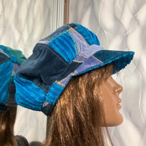 May include: A patchwork hat in shades of blue, teal, and purple. The hat features a corduroy texture and a small brim. The hat is made of various fabric squares sewn together with visible stitching. The hat is displayed on a mannequin head.
