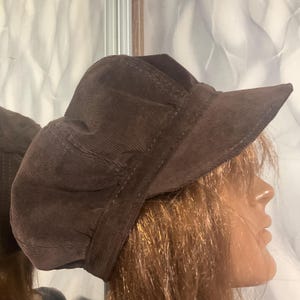 Puede incluir: Un sombrero de pana marrón con una visera corta. El sombrero tiene una copa redondeada y una banda en la base. La tela tiene una textura acanalada. El sombrero se muestra en una cabeza de maniquí.