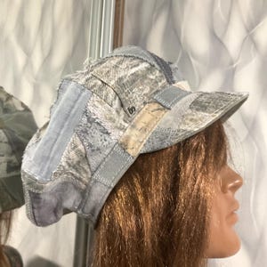 May include: A patchwork military-style cap constructed from various shades of light blue, grey, and white fabric panels. The hat features visible frayed edges, exposed stitching, and a structured brim, showcasing a multi-textured textile design.