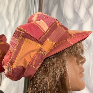 Puede incluir: Una gorra de patchwork confeccionada con varios paneles de tela en tonos rojos, rosas y naranjas. El gorro presenta costuras en zigzag amarillas visibles a lo largo de las uniones de la tela e incluye una visera rígida en la parte frontal.