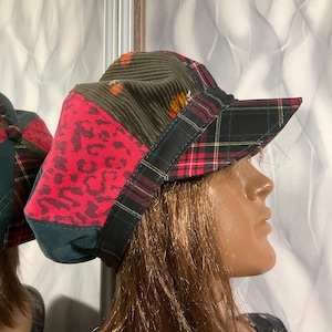 May include: A patchwork cap with a variety of patterns and textures. The cap incorporates red leopard print, olive green corduroy, and plaid fabric. The brim is plaid, and the cap has a black band. A unique fashion accessory.