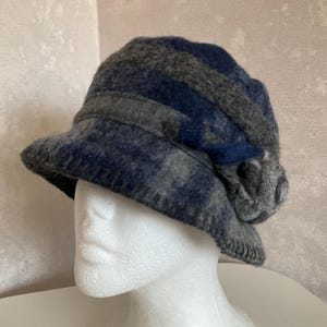 May include: A blue and grey cloche hat with a flower detail. The hat is made of a wool-like material and has a brim. The hat is displayed on a white mannequin head.