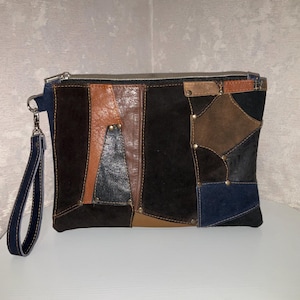 Puede incluir: Un bolso de mano patchwork con una correa de muñeca. La bolsa presenta una variedad de piezas de cuero en tonos marrón, negro y azul, cosidas juntas. Una cremallera plateada recorre la parte superior. La correa de muñeca es de mezclilla azul oscuro.