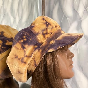 Puede incluir: Una gorra de estilo newsboy hecha de pana con un patrón tie-dye en tonos morado oscuro y amarillo dorado. El sombrero tiene una corona estructurada y una visera rígida y curva, mostrando una superficie de tela texturizada con un efecto moteado.
