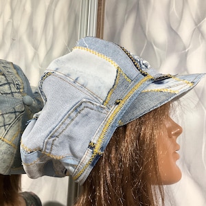 Puede incluir: Un sombrero de mezclilla patchwork con una visera curva y un diseño único. El sombrero está hecho de varios tonos de mezclilla azul, con costuras amarillas que añaden contraste. El sombrero se muestra en una cabeza de maniquí.