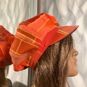 Puede incluir: Una gorra de patchwork en varios tonos de naranja y rojo, confeccionada con múltiples paneles de tela unidos mediante costuras en zigzag amarillas visibles. La gorra presenta una corona estructurada y una visera rígida con diseño texturizado.