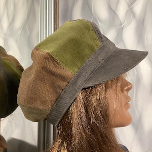 May include: A multi-coloured corduroy newsboy cap featuring panels of olive green, brown, and dark grey fabric. The hat has a structured dark grey brim and a wide band that encircles the base, showcasing a textured, ribbed corduroy material throughout.