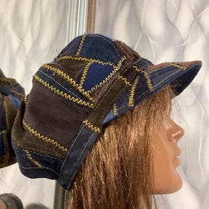Puede incluir: Gorra de estilo newsboy de patchwork en tonos azules, marrones y beige. La gorra presenta una variedad de telas cosidas con costuras amarillas. La visera es de color marrón liso. La gorra se muestra en una cabeza de maniquí.