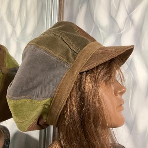 May include: A multi-colored corduroy cap featuring a patchwork design with panels in shades of olive green, grey, and brown. The cap has a structured brim and a textured fabric surface, constructed from several distinct sections of corduroy material.