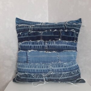 Op de afbeelding: Een blauwe denim kussenhoes gemaakt van gerecycled denim stof. De kussenhoes is gemaakt van meerdere stroken denim die aan elkaar zijn genaaid, waardoor een patchwork-effect ontstaat. De randen van de denim zijn gescheurd, waardoor de kussenhoes een rustieke uitstraling krijgt.