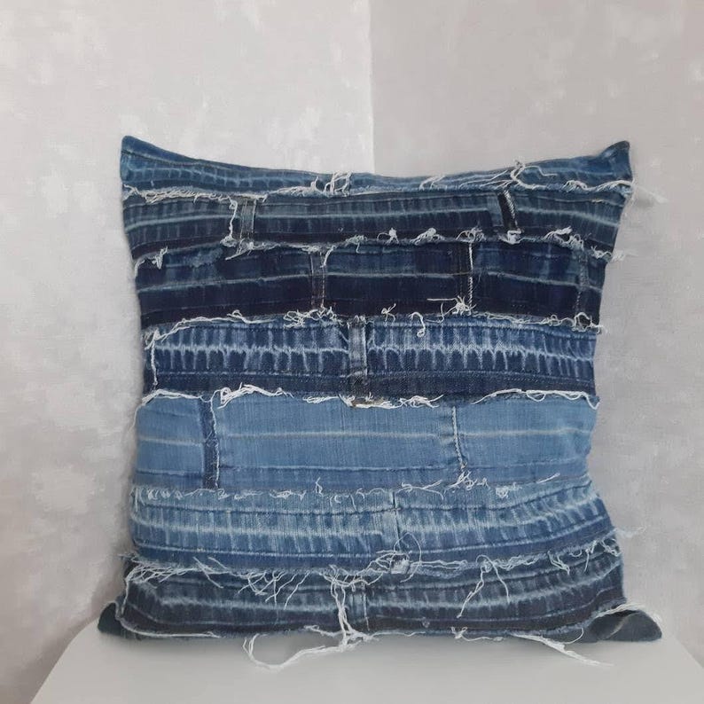 May include: A blue denim pillow cover made from repurposed denim fabric. The pillow cover is made of multiple strips of denim sewn together, creating a patchwork effect. The edges of the denim are frayed, giving the pillow cover a rustic look.