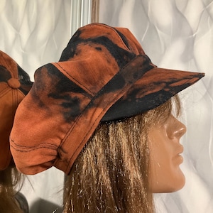 Puede incluir: Una gorra de estilo newsboy con un patrón tie-dye en color naranja óxido y negro. El sombrero tiene una corona estructurada con un botón central en la parte superior y una visera rígida y curva. La tela muestra una distribución irregular.
