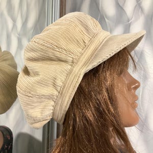 May include: A beige corduroy hat with a rounded crown and a small, stiff brim. The hat has a textured, ribbed appearance and a band around the crown. The hat is a neutral color and appears to be made of a soft fabric.