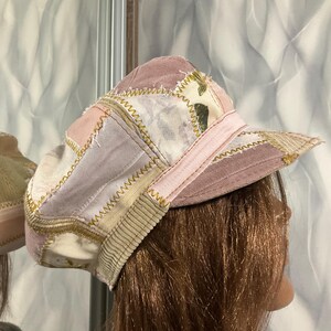 May include: A patchwork newsboy cap featuring various fabric panels in shades of dusty pink, cream, and beige. The cap is constructed with visible yellow zigzag stitching connecting the different textile sections and includes a structured brim.