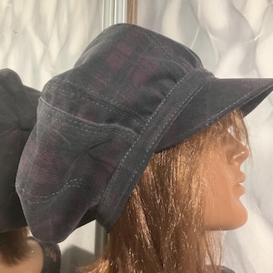 May include: A dark grey newsboy cap featuring a subtle, dark purple plaid pattern. The cap has a structured, rounded crown with multiple stitched panels and a stiff, curved brim extending from the front, providing a classic, full-coverage silhouette.