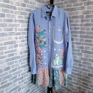 Könnte beinhalten: Ein hellblaues Leinenhemd mit Patchwork-Design mit floralen und geometrischen Mustern. Das Hemd hat einen Rüschen-Saum und einen weißen Spitzenbesatz.