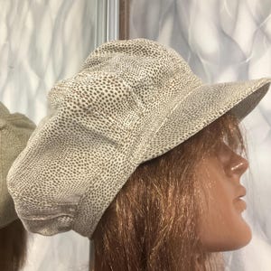 May include: A newsboy cap featuring a light beige base with a dense pattern of small, dark brown dots. The hat has a structured, rounded crown with stitched paneling and a stiff, curved brim extending from the front, providing a classic silhouette.