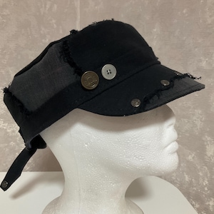 May include: A black military-style cap featuring distressed, frayed edges and a multi-panel construction. The hat is adorned with a circular bronze-toned button, a small light-coloured button, and several silver-toned metal studs along the brim.