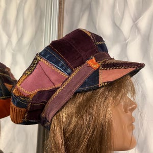 Könnte beinhalten: Eine Patchwork-Schiebermütze aus verschiedenen Stofftexturen wie dunkelviolettem Samt, blauem Denim und braunem Kord. Die einzelnen Stoffteile sind mit sichtbaren gelben Zickzacknähten verbunden und an der Seite befindet sich eine kleine orangefarbene Lasche.