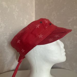 May include: A red military-style cadet cap featuring a repeating pattern of lighter red stars. The hat has a structured flat top, a short stiff brim, and two small dark metal grommets on the side. A fabric tie hangs from the back of the cap.