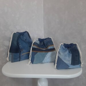 Può includere: Tre sacchetti a cordoncino in denim blu di diverse dimensioni, realizzati in tessuto denim riciclato. I sacchetti sono su un tavolo bianco.