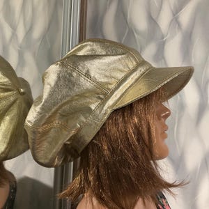 Puede incluir: Una gorra de estilo newsboy en color dorado metálico con visera estructurada y diseño de corona de varios paneles. El sombrero tiene una superficie lisa y reflectante con costuras visibles, creando una silueta redondeada clásica.